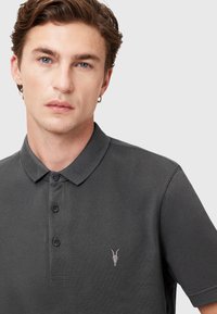 Graues Poloshirt aus strukturiertem Stoff, mit einer Knopfleiste mit drei Knöpfen und einem kleinen gestickten Logo auf der linken Brust.