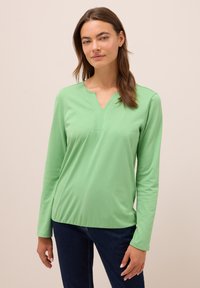 Frau mit langen braunen Haaren, die ein hellgrünes langärmliges V-Ausschnitt-Shirt und dunkelblaue Jeans trägt, steht vor einem schlichten hellen Hintergrund.