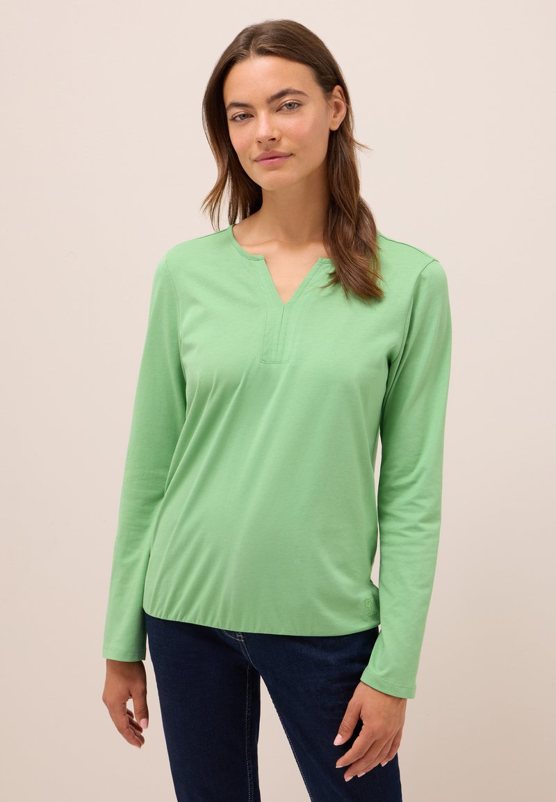 Frau mit langen braunen Haaren, die ein hellgrünes langärmliges V-Ausschnitt-Shirt und dunkelblaue Jeans trägt, steht vor einem schlichten hellen Hintergrund.