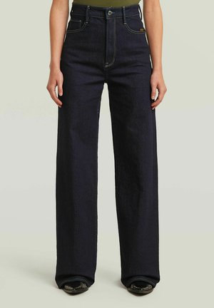 Slim-waist high-rise dark blue wide-leg jeans worn with black shoes and a green tucked-in top.