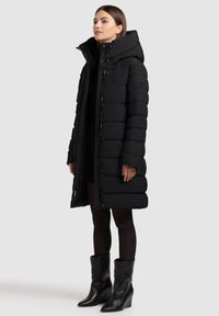 Černý péřový kabát s vysokým límcem, dlouhými rukávy a lesklým povrchem. Má zapínání na knoflíky a zip, stylizovaný s vysokými černými botami.