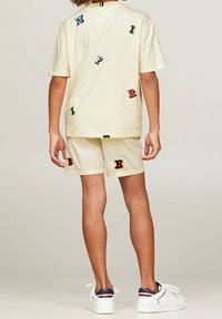 Sæt bestående af en gul bomulds skjorte og shorts, der har spredte teksturerede bogstav-applikationer i grøn, rød, sort og orange. Hvide sneakers fuldender looket.