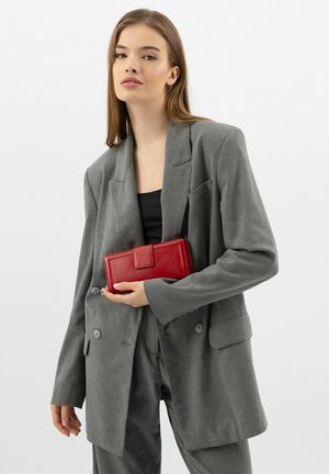 Rote Lederbrieftasche, die in den Händen gehalten wird, mit einem Klappenverschluss. Tragen zu einem grauen Blazer und einem schwarzen Oberteil. Einfaches, strukturiertes Design.