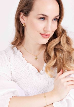 Bijoux en or avec un pendentif rond et une chaîne délicate, portés avec un chemisier blanc texturé, mettant en valeur des ondulations douces dans les cheveux.