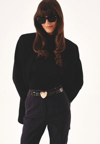 Blazer negro oversize sobre un cuello de tortuga negro, combinado con pantalones de mezclilla oscuros. Presenta una brillante hebilla de cinturón en forma de corazón dorado. Gafas de sol negras.