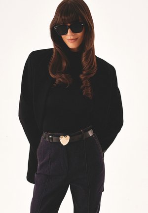 Schwarzer übergroßer Blazer über einem schwarzen Rollkragenpullover, kombiniert mit dunklen Jeans. Ausgestattet mit einer glänzenden goldenen herzförmigen Gürtelschnalle. Schwarze Sonnenbrille.