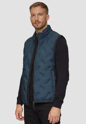 Gepolsterte türkisfarbene Weste mit einem hohen Kragen und Reißverschluss, kombiniert mit einem schwarzen Langarmshirt. Die Weste hat eine gesteppten Textur.