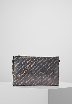 Pochette rectangulaire noire avec un imprimé du logo Versace Jeans en diagonale et une bandoulière chaîne dorée reposant sur un socle blanc.