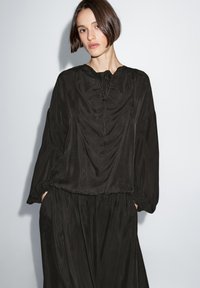Schwarzes Kleid mit drapiertem Design, runder Halslinie, langen Ärmeln und elastischen Manschetten. Hergestellt aus einem glatten, leichten Stoff.
