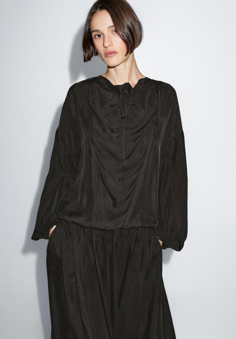 Schwarzes Kleid mit drapiertem Design, runder Halslinie, langen Ärmeln und elastischen Manschetten. Hergestellt aus einem glatten, leichten Stoff.