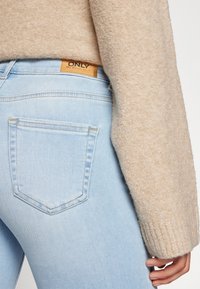 Vaqueros de corte slim en azul claro, con dos bolsillos traseros y una etiqueta de marca de cuero marrón en la cinturilla. Manga de un suéter texturizado en beige visible.