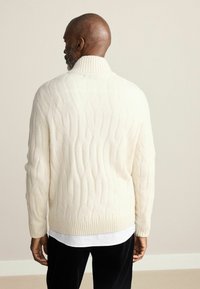 Cream cable-knit sweater with a ribbed collar and hem, featuring a textured pattern, worn over a white shirt and paired with dark pants.