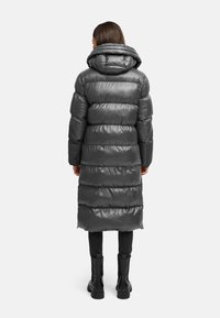 Long manteau rembourré gris foncé avec un col haut et des coutures.horizontal. Le matériau semble brillant et rembourré ; porté sur des leggings noirs et des bottes.