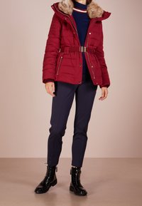 Punainen tikattu takki, jossa tekoturkistakki, vyötäröllä vyö ja hopeiset yksityiskohdat, yhdistetty tummansinisiin housuihin ja mustiin nyörikenkiin.