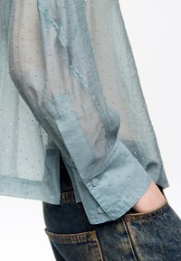 Jasnoniebieska przezroczysta koszula z małymi srebrnymi ćwiekami, z mankietami na guziki i luźnym krojem, w połączeniu z ciemnoniebieskimi dżinsami.