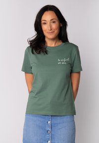 T-shirt vert à manches courtes avec un col rond et un texte blanc indiquant "So einfach ist das," associé à une jupe en denim.