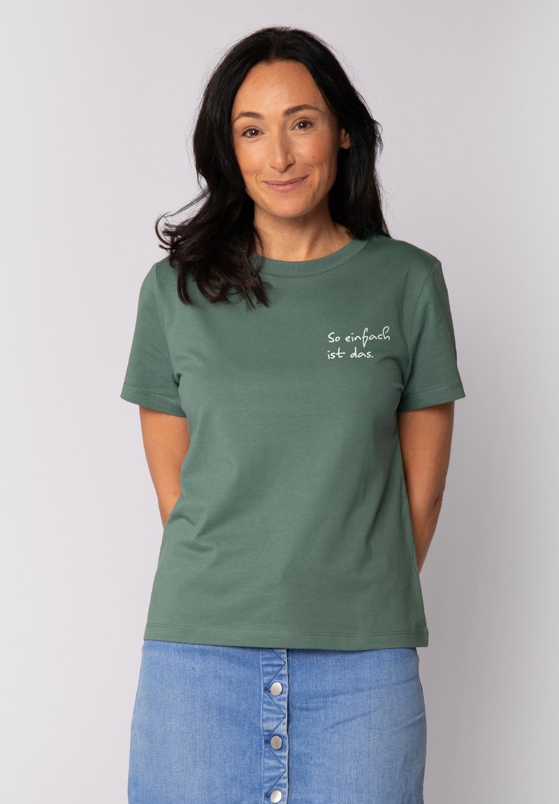 T-shirt vert à manches courtes avec un col rond et un texte blanc indiquant "So einfach ist das," associé à une jupe en denim.
