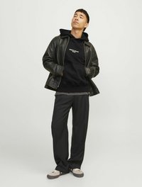 Jachetă din piele neagră peste un hanorac negru cu text alb, tricou gri, pantaloni negri largi și pantofi bej cu talpă închisă. Fundal neutru.