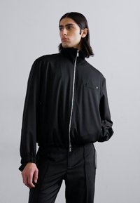 Black zip-up jacket with high collar, elastic cuffs, and a chest pocket; made from lightweight, smooth fabric. Paired with dark trousers.