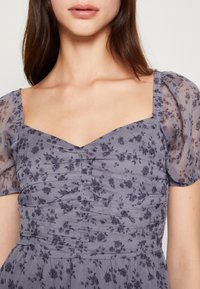 Žena oblečená v světle fialových šatech s průhlednými puff rukávy a malým tmavým květinovým vzorem, odhalující dekolt a horní část trupu.