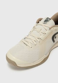 Chaussure de sport beige avec une tige en mesh, surface texturée et semelle en caoutchouc. Présente des lacets et un logo sur le côté.