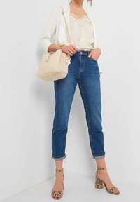 Beige axelremsväska, krämfärgad topp med spetsdetaljer, vit kofta, mörkblå uppvikta jeans och högklackade sandaler med ormskinnsmönster.