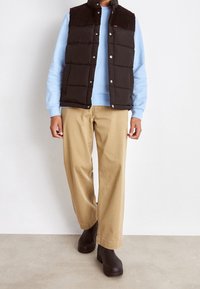 Chaleco acolchado negro con cuello de pana sobre una sudadera azul claro, combinado con pantalones anchos beige y zuecos negros.