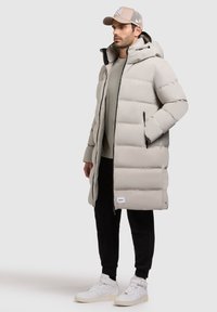 Lang, lys grå dunjakke med hætte, med lynlås foran, to sidelommer og et logo-patch forneden. Bæres med sorte bukser og hvide sneakers.