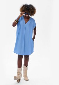 Jasnoniebieska krótka sukienka z dekoltem w kształcie litery V i luźnym krojem, z bocznymi kieszeniami. Noszona z beżowymi kowbojkami i dużymi okularami przeciwsłonecznymi.