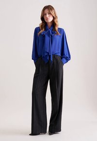 Blusa blu con un grande fiocco al collo, abbinata a pantaloni neri a vita alta e gamba larga con righe verticali bianche. Tessuto dalla texture liscia.