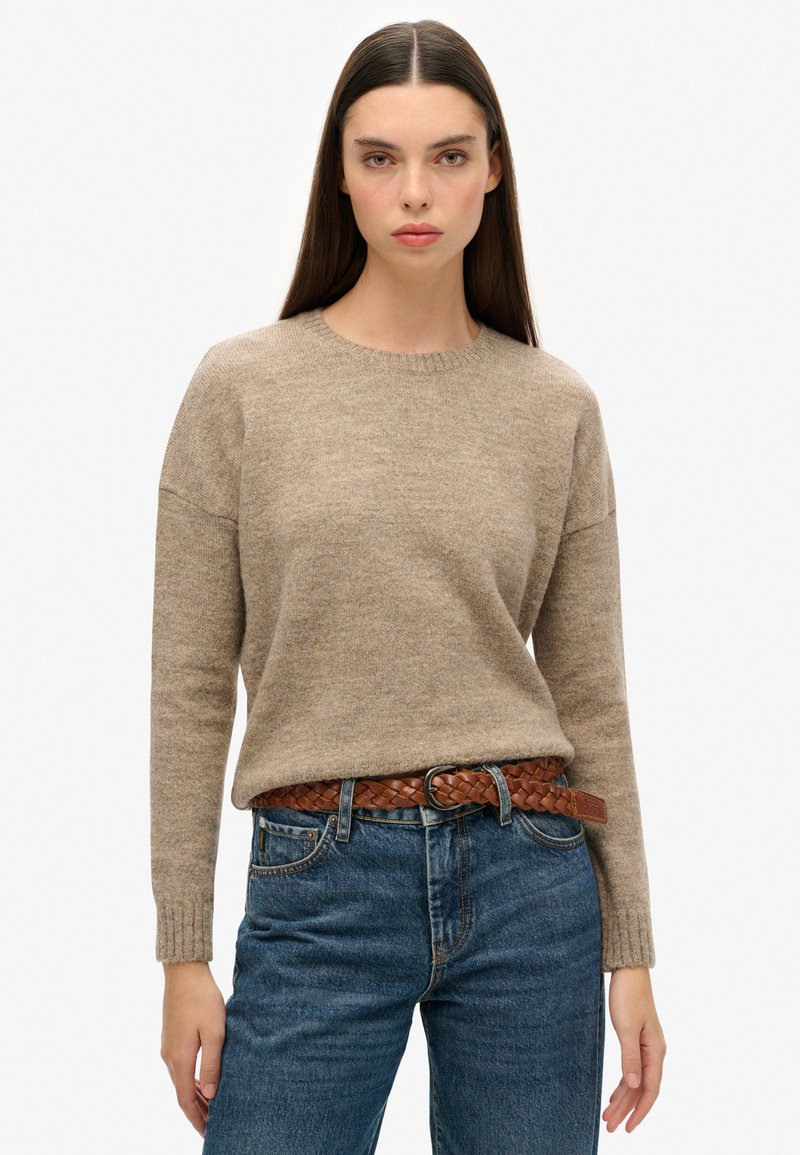 Pull en tricot beige avec un col rond et des poignets côtelés, associé à un jean en denim bleu foncé et une ceinture en cuir tressé marron.