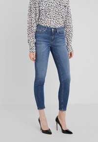 Mujer vistiendo jeans ajustados azules, tacones altos negros y una blusa blanca con manchas abstractas negras, de pie frente a un fondo gris claro liso.