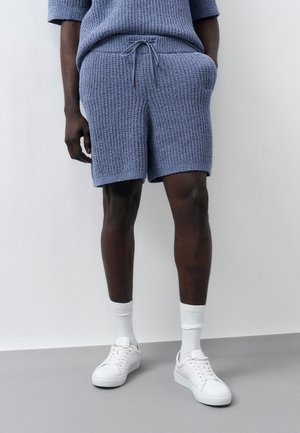 Osoba nosiaca textúrované modré pletené šortky a zodpovedajúci top, stojaca s rukami v kapsách, biele ponožky a biele tenisky na šedom podlahe.