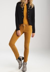 Czarna krótka kurtka z srebrnymi guzikami, żółty sweter w prążki, musztardowe rurki oraz czarno-białe botki w panterkę.
