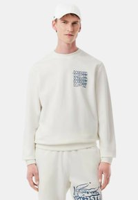 Sudadera blanca con puños y dobladillo de canalé, presenta una impresión del logo azul de Lacoste en el bolsillo del pecho, combinada con pantalones de chándal a juego.