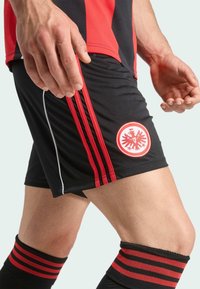 Sorte atletiske shorts med røde striber, der har et rundt broderet emblem og en komfortabel pasform, parret med ribbede sorte og røde sokker.