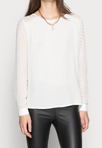 Blouse blanche avec des manches transparentes texturées arborant un motif rayé, encolure ronde et poignets retroussés, associée à un pantalon noir ajusté.