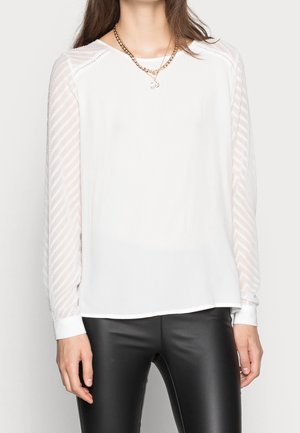 Blouse blanche avec des manches transparentes texturées arborant un motif rayé, encolure ronde et poignets retroussés, associée à un pantalon noir ajusté.