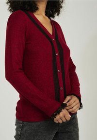 Czerwony kardigan z długim rękawem, z dekoltem w serek, czarnymi paskami na środku i koronkowym wykończeniem przy mankietach; posiada srebrne guziki z przodu.