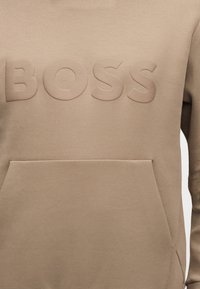 Μπεζ φούτερ με μεγάλους ανάγλυφους χαρακτήρες που σχηματίζουν τη λέξη "BOSS" στο στήθος και μια μπροστινή τσέπη καγκουρό.