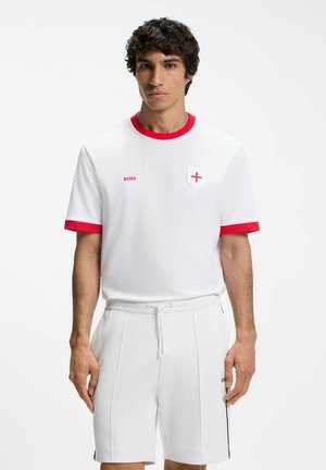 Jonge man die witte sportkleding draagt met rode bies en een klein rood kruis op de borst, staand tegen een effen witte achtergrond.