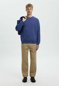 Mėlynas džemperis su apykakle, smėlio spalvos plačios kelnės ir juodi batai. Modelis laiko juodą rankinę. Paprastas, kasdieniškas apranga.