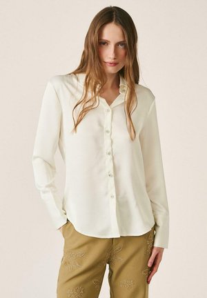 Mujer con cabello largo y castaño, vistiendo una blusa abotonada de color blanco roto y pantalones beige con bordado floral, de pie contra un fondo liso.