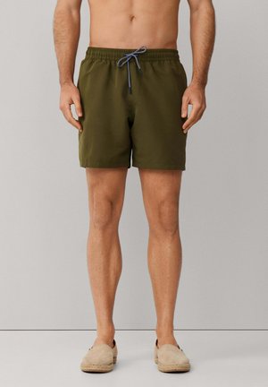 Man in olijfgroene zwembroek met zwart-witte trekkoord en beige instappers, staand tegen een effen achtergrond.