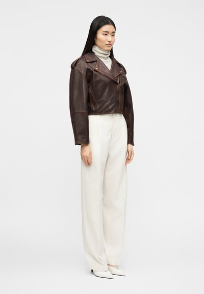 Νέα γυναίκα που φορά καφέ δερμάτινο μπουφάν, ριγέ πουλόβερ με λαιμόκοψη, φαρδιά λευκά παντελόνια και λευκά ψηλοτάκουνες μυτερές γόβες, στέκεται μπροστά σε λευκό φόντο.