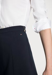 Laivastonsiniset housut ovat muotoiltuja ja niiden pinta on sileä. Niissä on hopeinen nappi ja pieni logo-patch punaisena, valkoisena ja sinisenä.