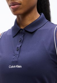Κοντινό πλάνο ενός ατόμου που φοράει μπλε σκούρο αμάνικο πόλο Calvin Klein με γιακά με κουμπιά και λευκό λογότυπο στο στήθος.