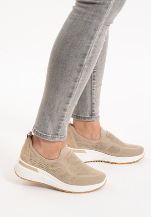 Persona que lleva vaqueros ajustados gris claro y zapatillas tipo slip-on de punto beige con suelas blancas, de pie sobre una superficie blanca.