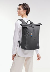 Sac à dos gris à rabat fabriqué en tissu avec une surface texturée, comprenant une poche zippée sur le côté et un accent coloré sur le tirant de la fermeture éclair.