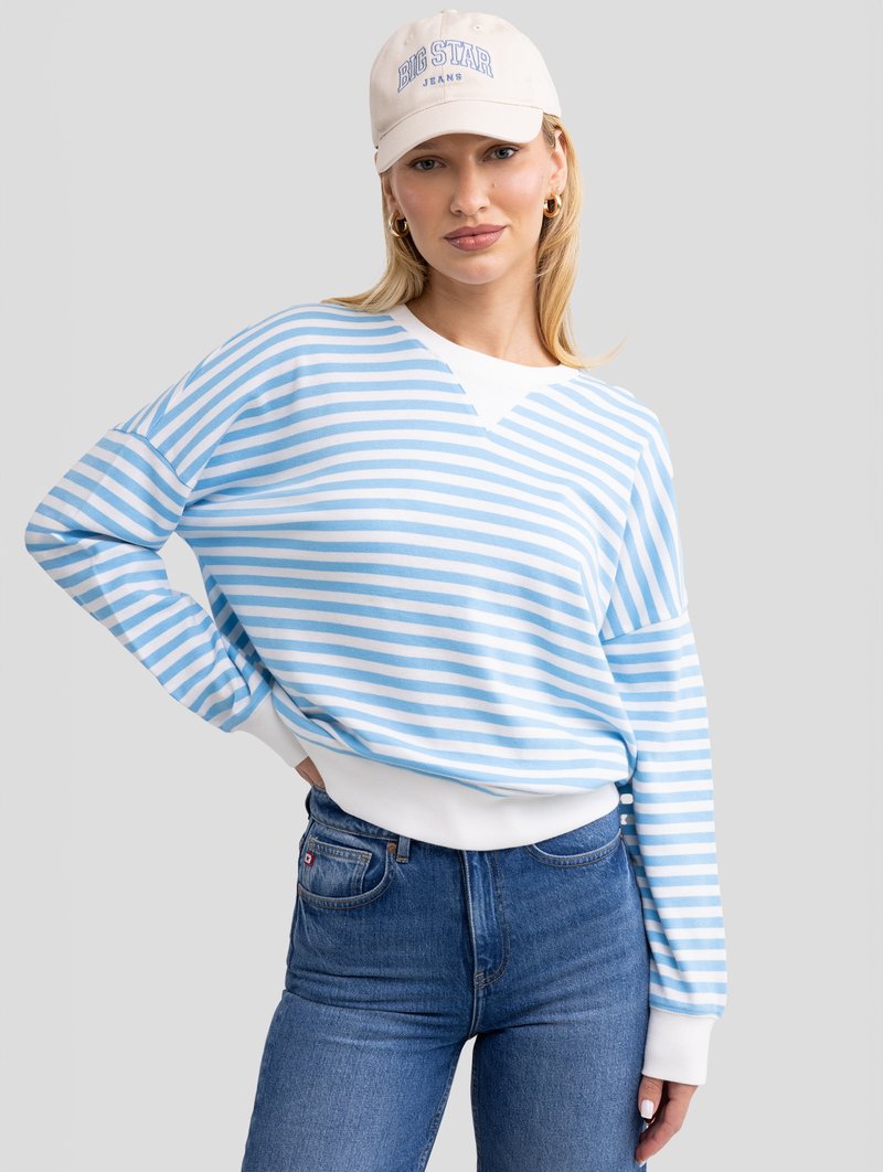Sweat-shirt rayé bleu et blanc avec un col en V, poignets blancs ajustés, associé à un jean bleu. Le mannequin porte une casquette beige avec un logo.